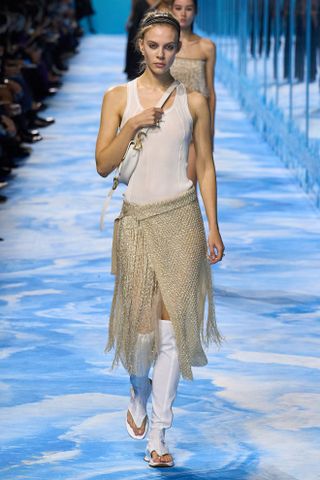 Model at dior wearing a white tank, gold fringe skirt, white thong sandals, and a white crossbody bag.