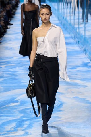 A woman wears a white one shoulder button-down shirt, black midi skirt, black bag, and black knee high shoes.