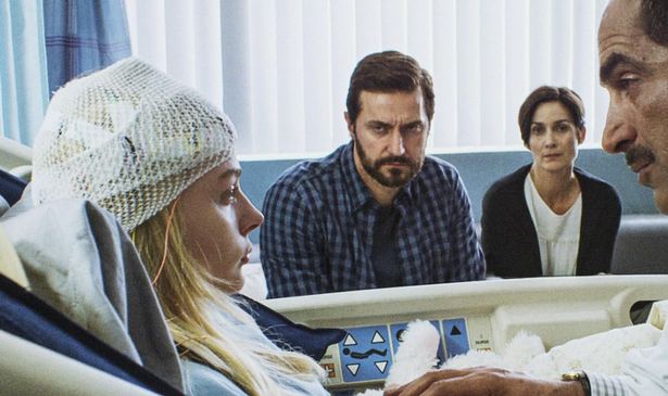 A group of people are sat around a woman in a hospital bed