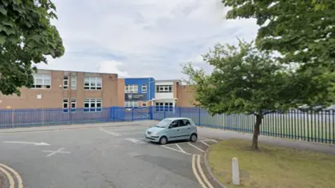 Google A screenshot from Google Streetview of a sprawling brick building with a small carpark out the front with a small blue car parked in it.  The building is surrounded by a blue fence and a couple of trees.