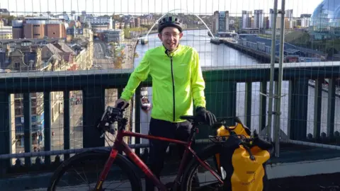 Chris Willian A man holding his cycle bike on a bridge with the river behind him, on either side and roads and buildings and clear skies behind.
