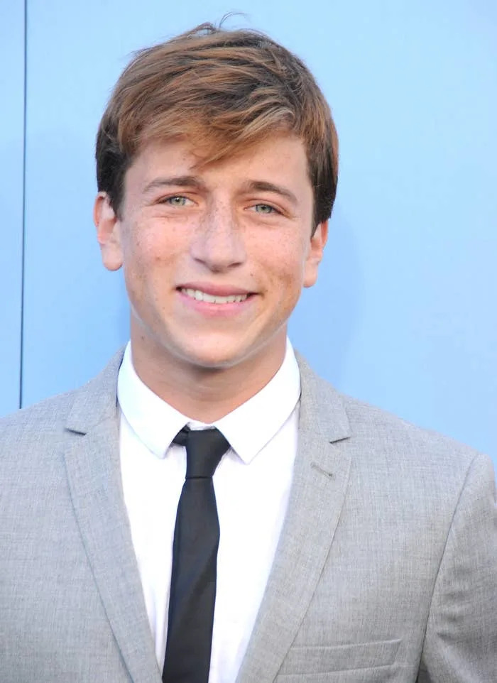 Skyler Gisondo in a gray suit with a black tie smiles at the camera against a plain backdrop