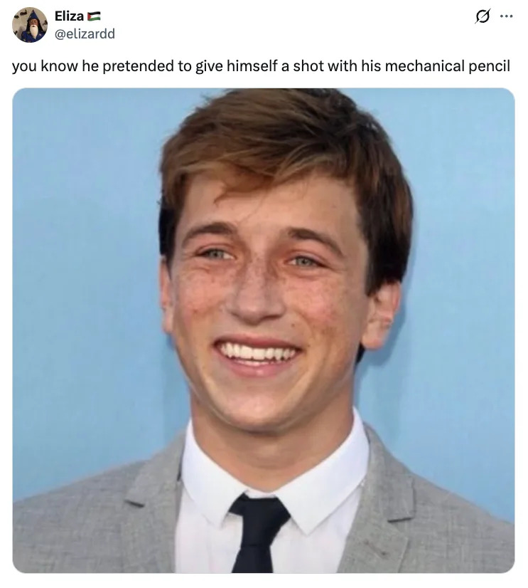 Skyler Gisondo smiling in a gray suit and black tie against a plain backdrop. Caption jokes about pretending to give himself an injection with a mechanical pencil