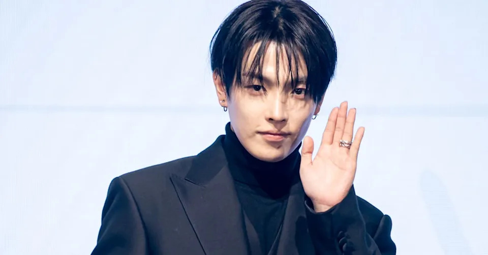 ATEEZ's Hongjoong with dark hair wearing a black suit and turtleneck, raising their hand in a waving gesture.