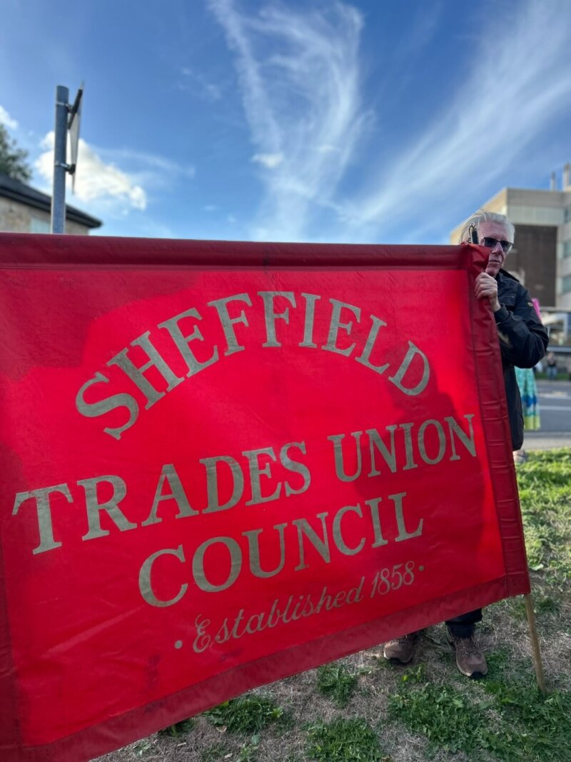 A photo of a banner at a protest with the words "SHEFFIELD TRADES COUNCIL" on it, and a man standing next to it