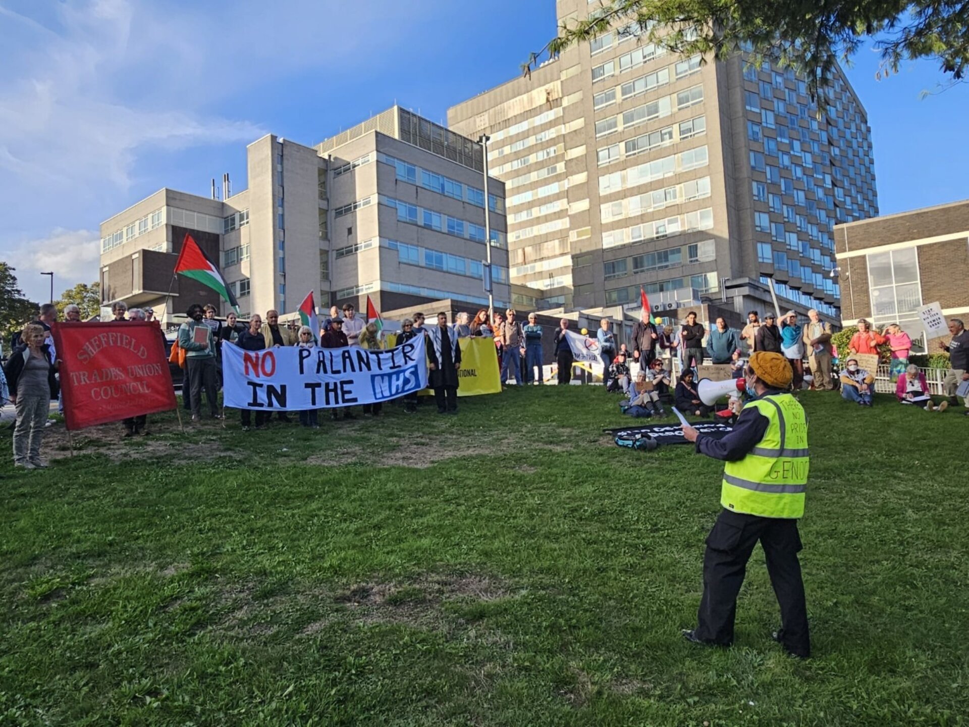 A photo of a man in high-viz addressing a demonstration throigh a loudspeaker, with people holding banners and listening to him