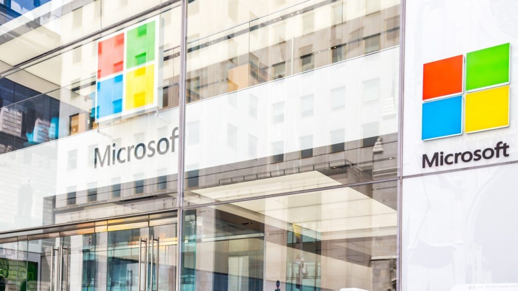 The Safest Upside in the Mag 7 Right Now The modern glass facade of a Microsoft building, featuring the iconic four-square logo in red, green, blue, and yellow, and the 'Microsoft' wordmark. Reflections of other buildings and the sky are visible on the polished glass, with the logo repeated on the interior through the glass.