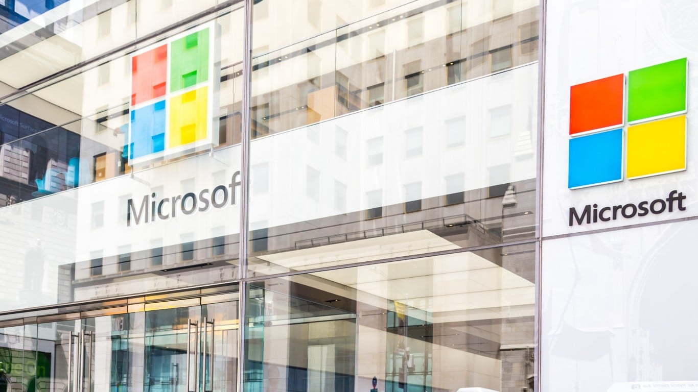 The Safest Upside in the Mag 7 Right Now The modern glass facade of a Microsoft building, featuring the iconic four-square logo in red, green, blue, and yellow, and the 'Microsoft' wordmark. Reflections of other buildings and the sky are visible on the polished glass, with the logo repeated on the interior through the glass.