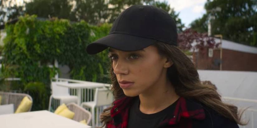 Hannah John-Kamen wearing a baseball cap and staring ahead on an outdoor patio in The Stranger.