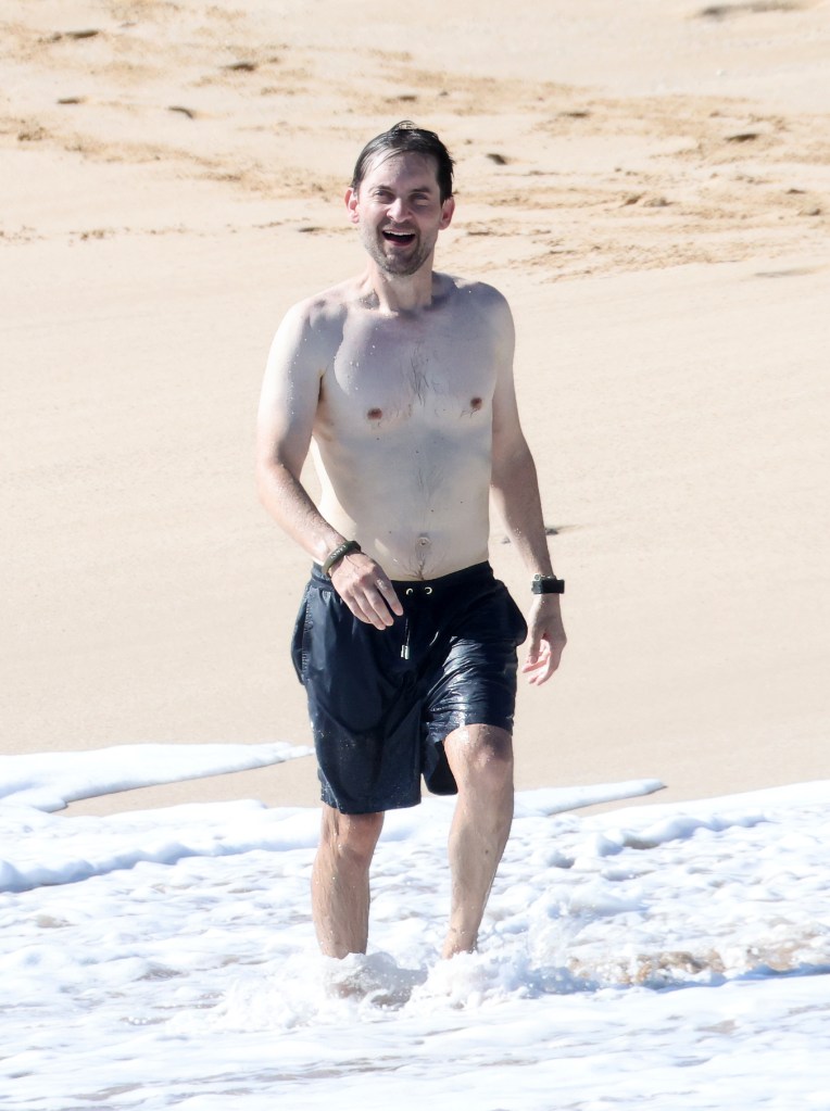 Tobey Maguire smiling and walking out of the ocean at a beach in Hawaii.