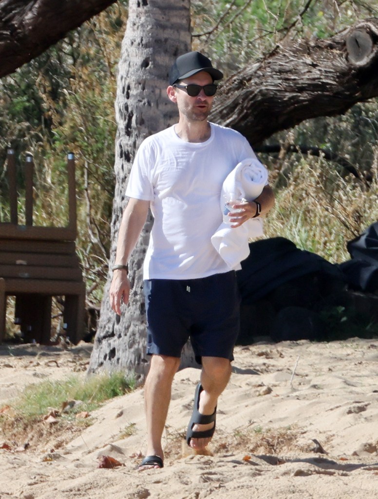 Tobey Maguire walking on a sandy beach in Hawaii, wearing a white t-shirt, dark shorts, a black cap, and sunglasses, while holding a towel.