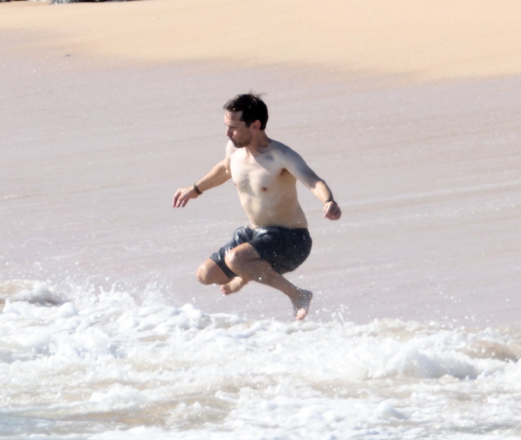 Tobey Maguire in black swim trunks jumps over waves on a beach in Hawaii.