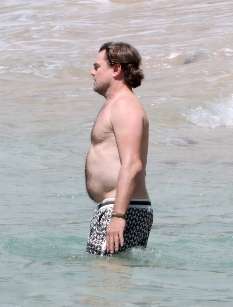 Leonardo DiCaprio stands shirtless in the ocean, wearing patterned swim trunks during a beach getaway in Hawaii.