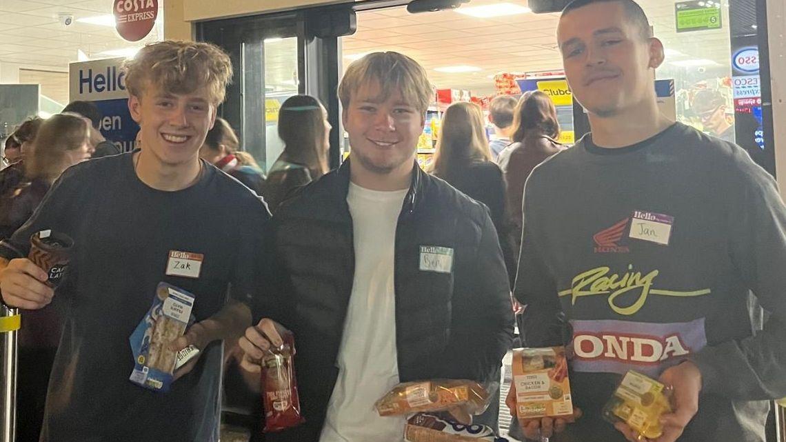 Three young men stand in front of a Tesco and hold up their meal deals.