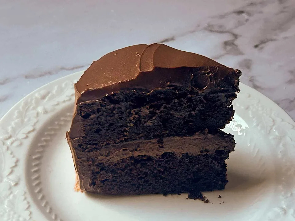 Chocolate layer cake slice with rich frosting on a decorative white plate.