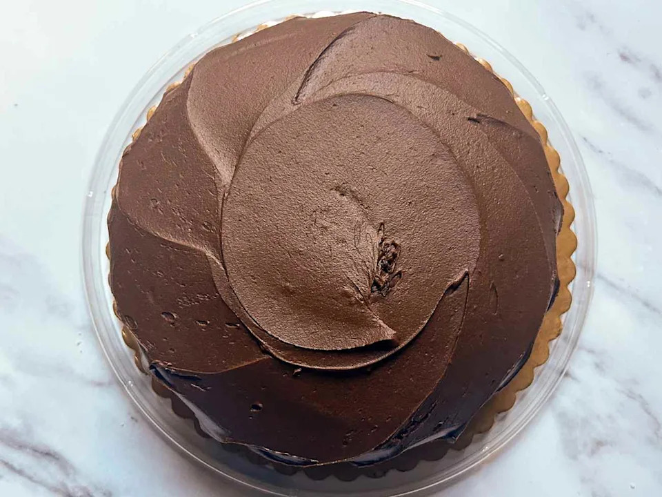 Chocolate frosted cake with smooth icing, displayed in a clear plastic container on a marble surface.