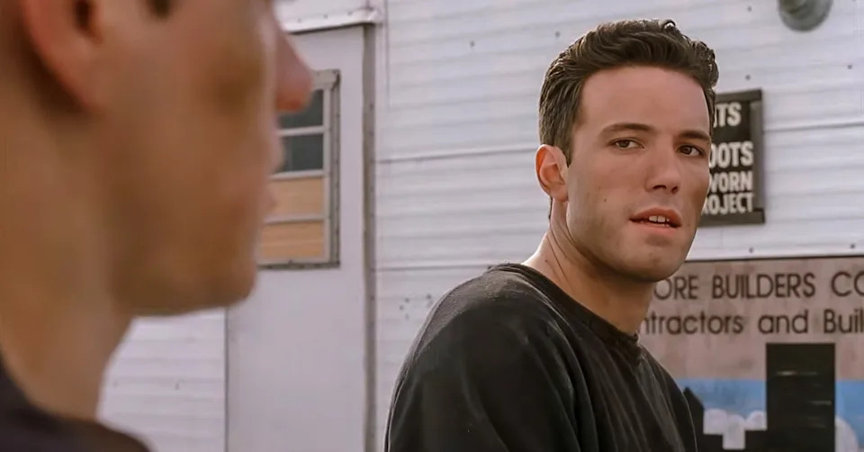 Two people are conversing outdoors near buildings. One faces away, while the other looks intently at them