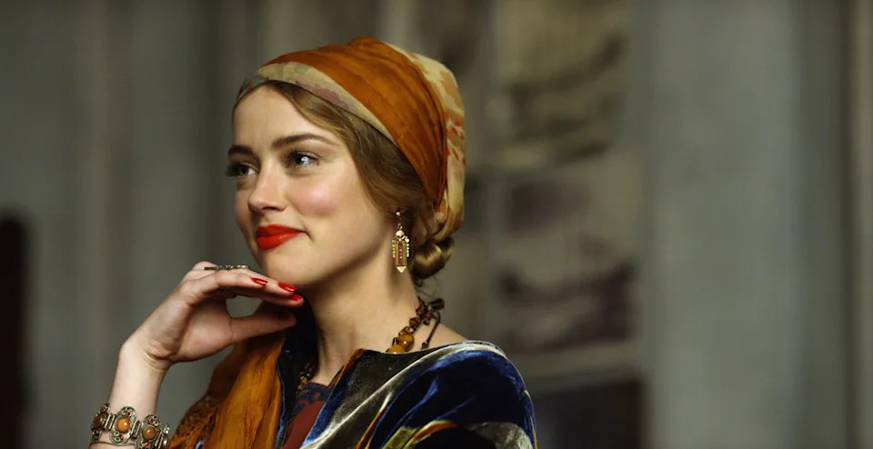 Person wearing a patterned headscarf and earrings, posed with a thoughtful expression, hand near chin