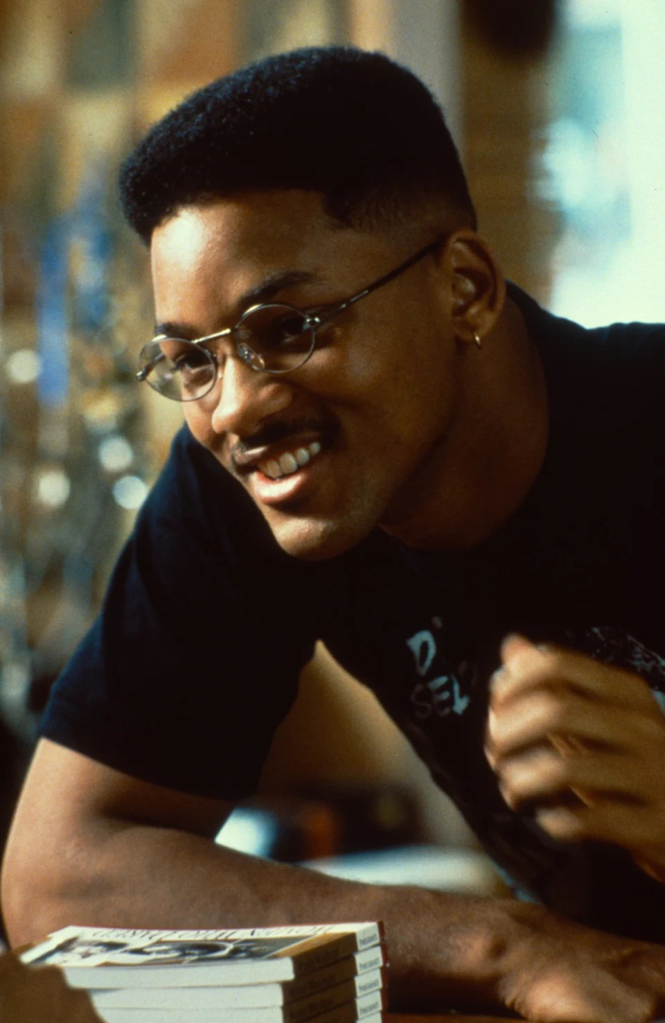 Person with a high-top fade hairstyle, wearing glasses and a casual t-shirt, smiling and leaning forward near a stack of books