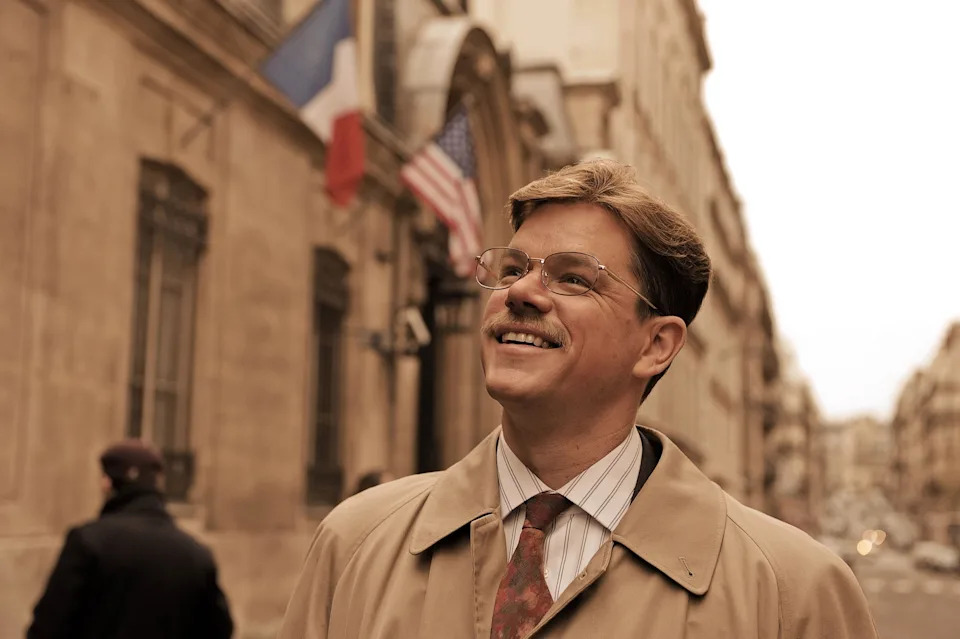A person in a trench coat and tie smiles while standing on a street with French and American flags in the background