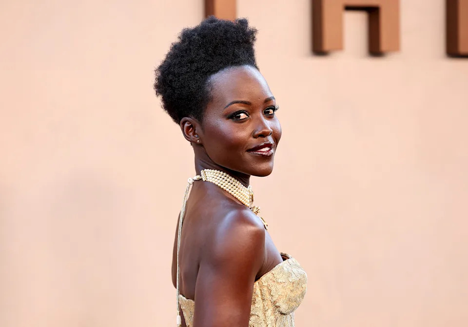 Person in elegant strapless dress, adorned with pearls, poses on a red carpet, exuding confidence