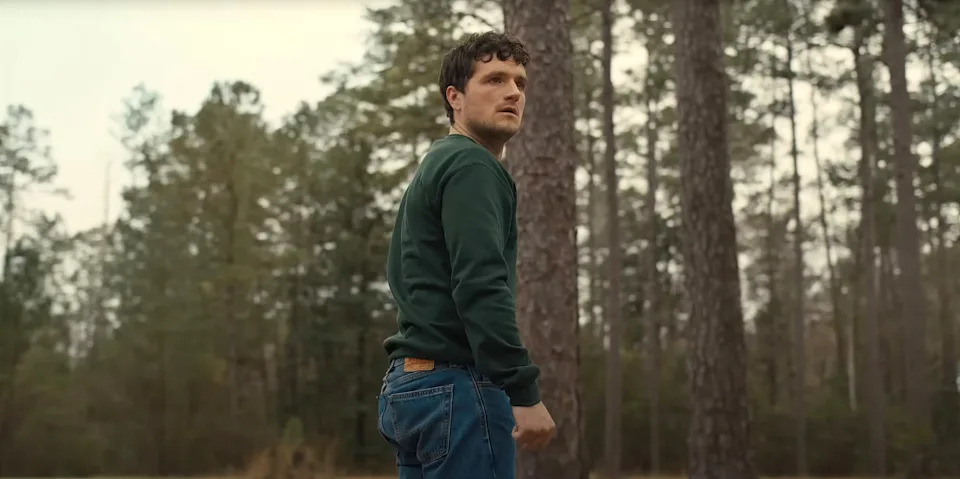 A person stands in a wooded area, wearing a casual green sweater and jeans, looking intently into the distance