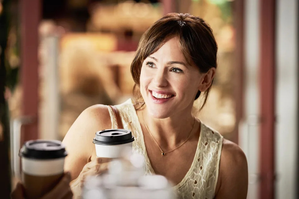 Person smiling while holding a takeaway coffee cup, wearing a sleeveless lace top, seated indoors