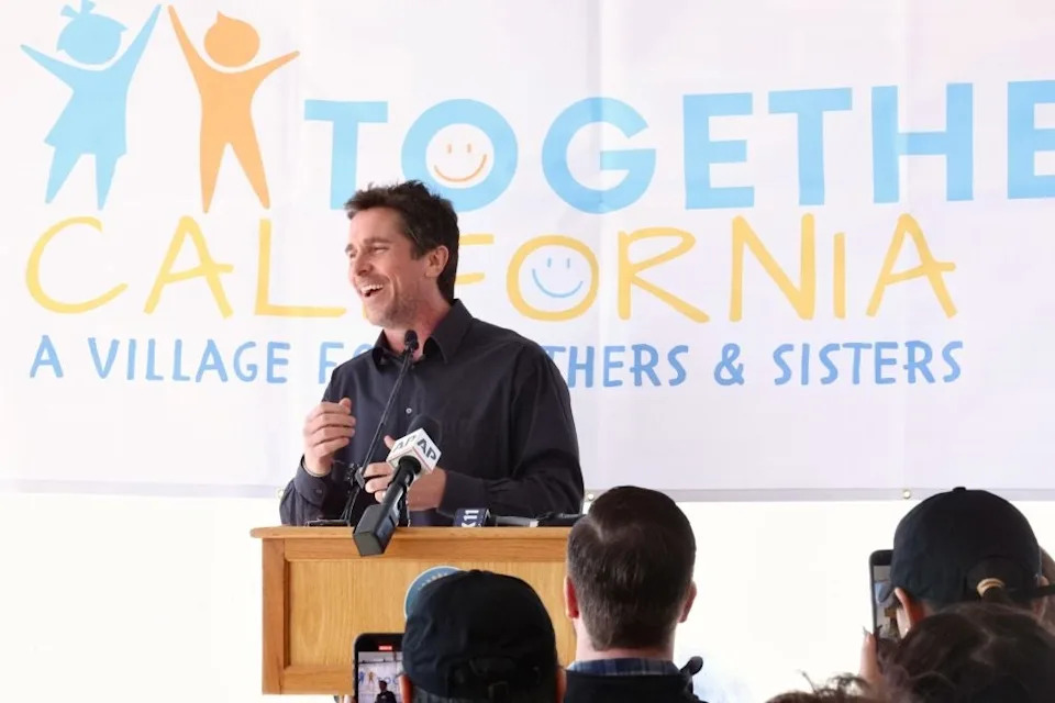 Man speaking at a podium with a "Together California" banner in the background. Audience members are taking photos