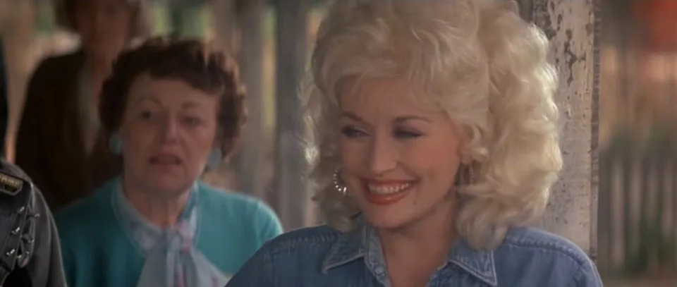 Person with curly hair smiling in a denim outfit, standing outdoors with another person in the background