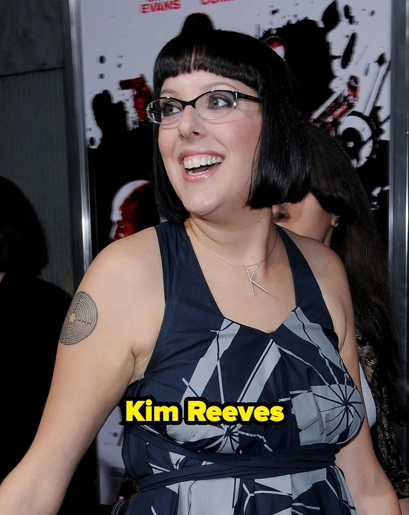 Person with short hair, wearing a patterned sleeveless dress and glasses, smiling at a formal event