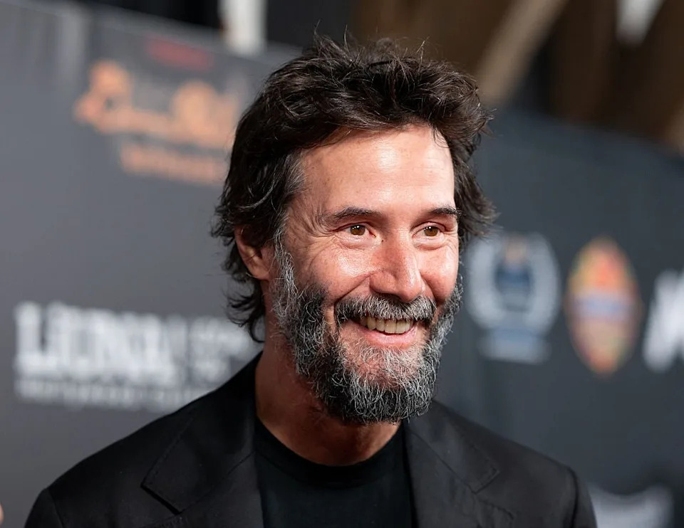 A person with dark hair and a beard smiles at an event, wearing a black blazer over a black shirt