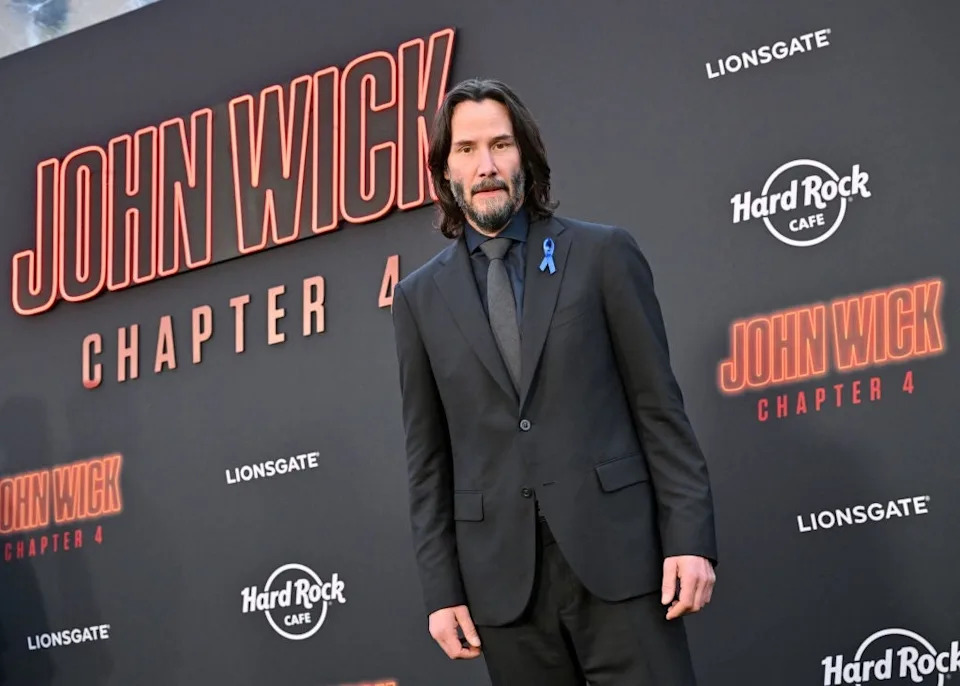Person in a tailored suit at a movie premiere event, standing in front of a backdrop with "John Wick: Chapter 4" text