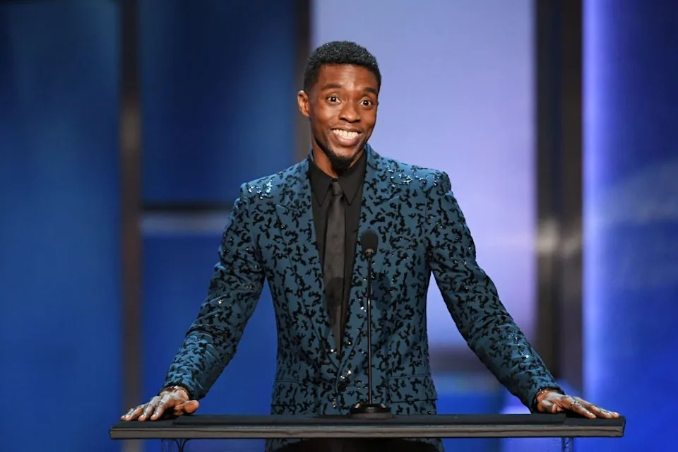 A man in a patterned suit smiles while speaking at a podium during an event