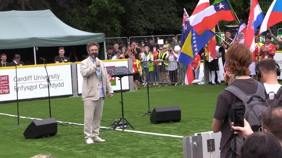 Man in a light suit speaks on a green field at an outdoor event with audience holding various national flags