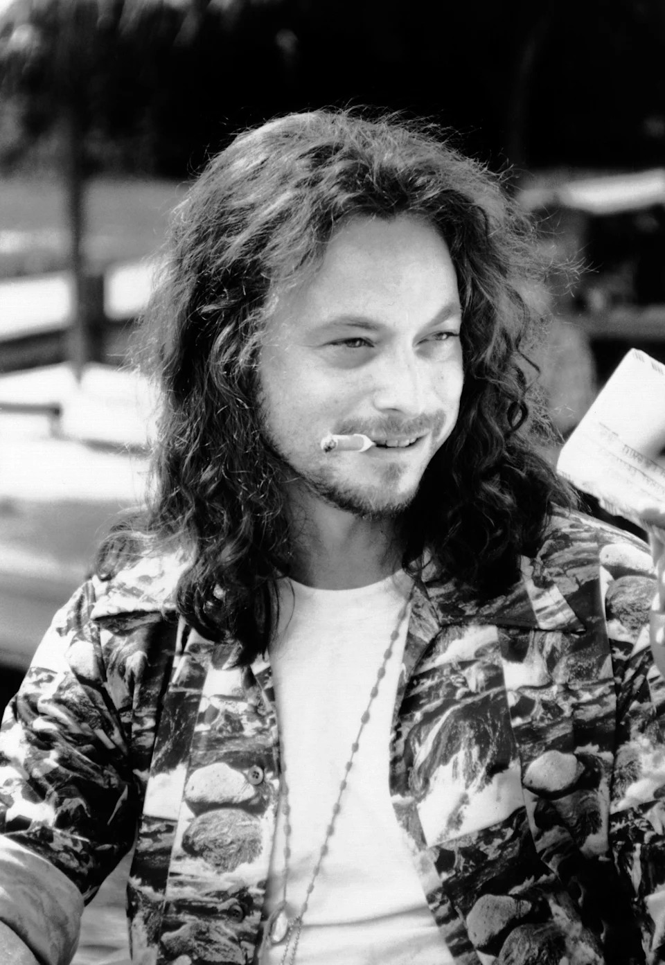Person with long hair smiles while holding a cigarette. They wear a patterned shirt over a plain T-shirt in a casual outdoor setting