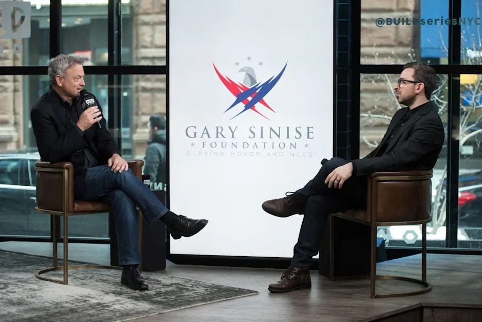 Two people sit and talk on stage at an event for the Gary Sinise Foundation; one holds a microphone
