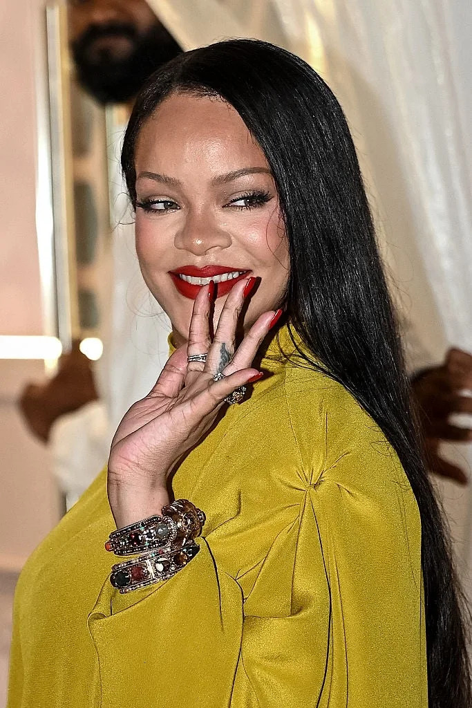 Person with long hair wearing elegant attire, smiling with hand near face, displaying jewelry