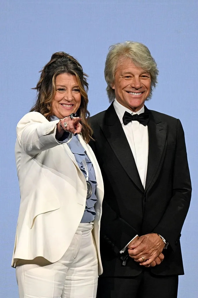 A woman in a white suit points forward while standing beside a man in a black tuxedo, both smiling against a plain background
