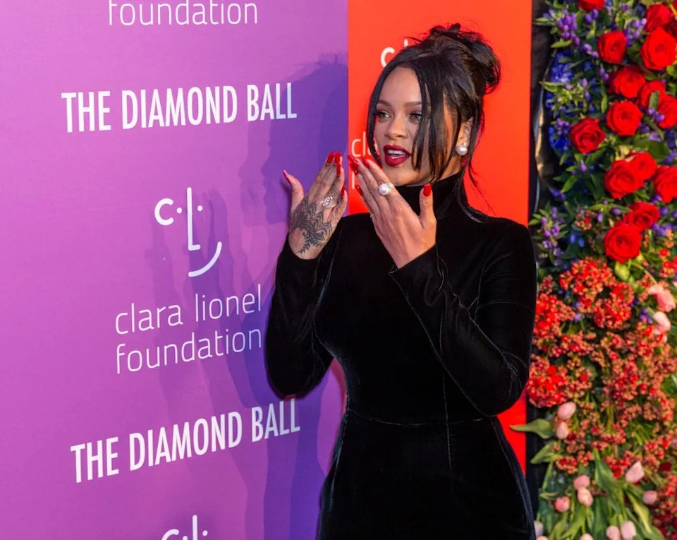 Person in elegant velvet outfit at a red carpet event, with flowers and foundation logo in background