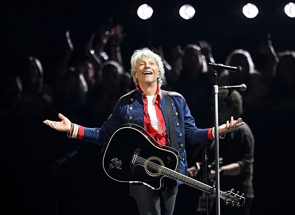 Musician performs energetically on stage with a guitar, wearing a jacket with decorative buttons. Audience claps in the background