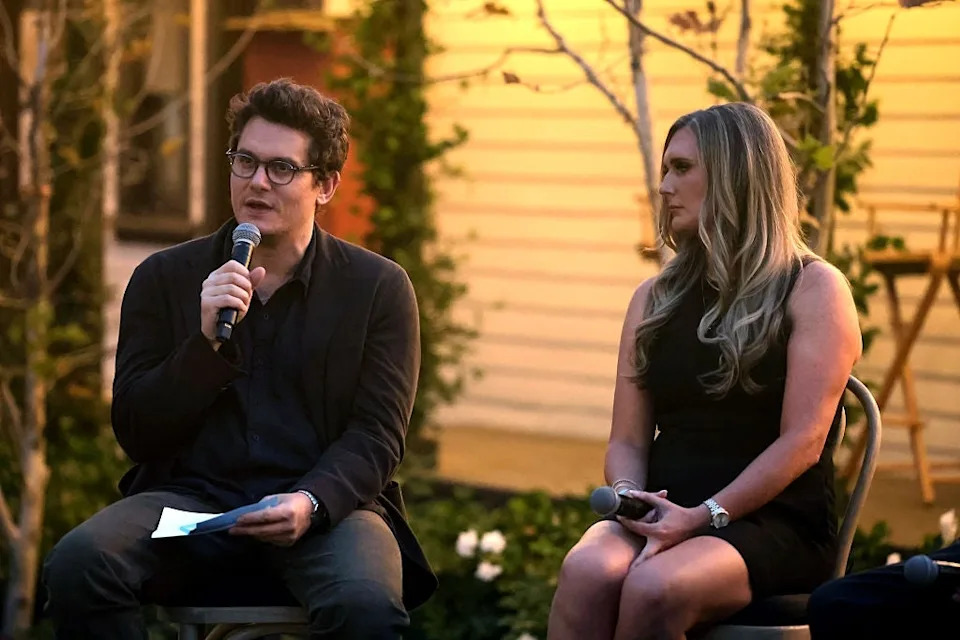 A man and woman sit outside, speaking into microphones. The man wears a dark suit, and the woman wears a sleeveless dress