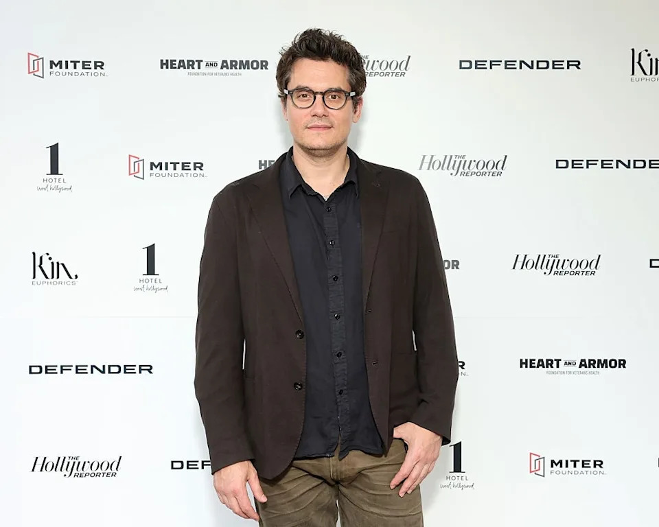 Person in glasses wearing a dark suit and shirt at a media event backdrop featuring logos, including "Hollywood Reporter" and "Heart and Armor."