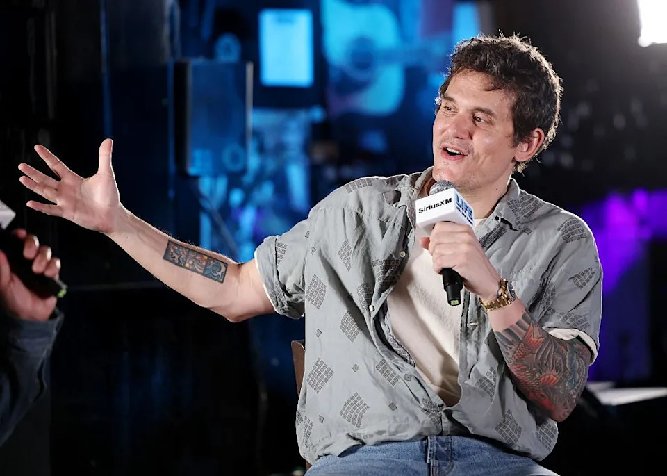 Person wearing casual patterned shirt and jeans, holding a microphone, gesturing enthusiastically during an interview