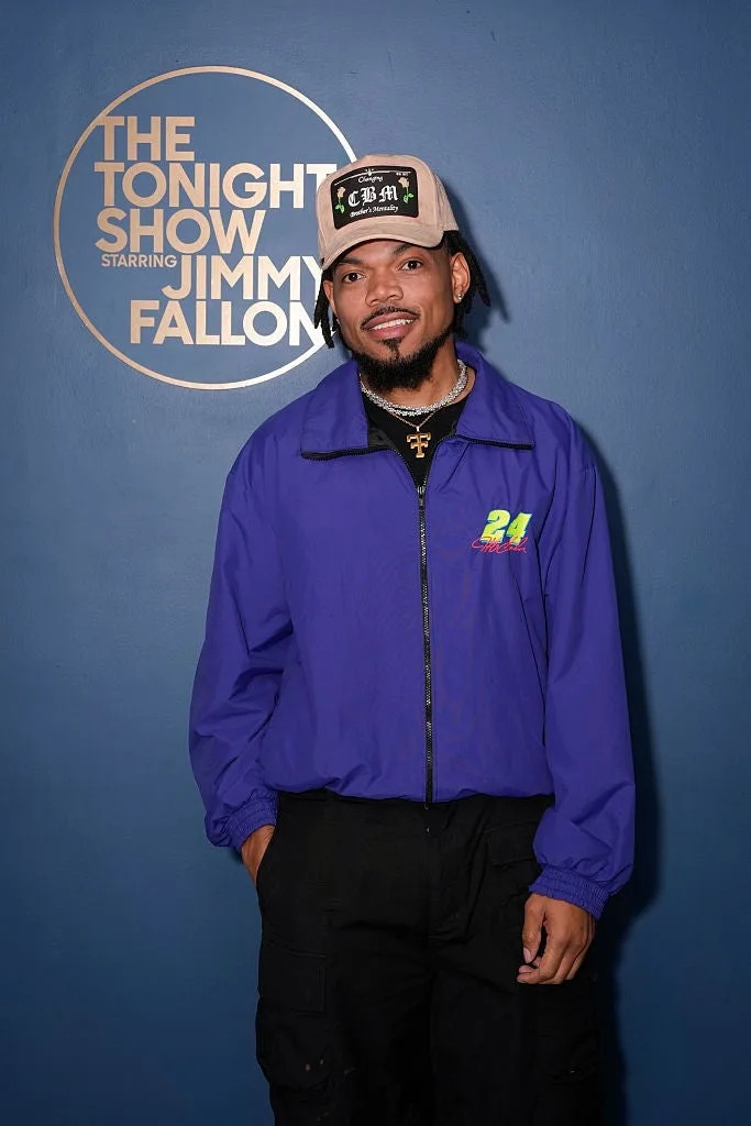 A person wearing a casual cap, blue jacket, and black pants poses with a smile in front of "The Tonight Show Starring Jimmy Fallon" backdrop