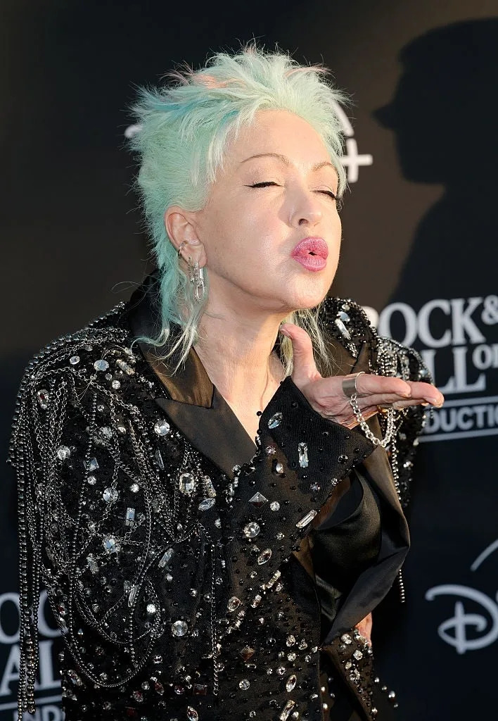 Person with short, spiky hair in a sequin jacket blows a kiss on a red carpet