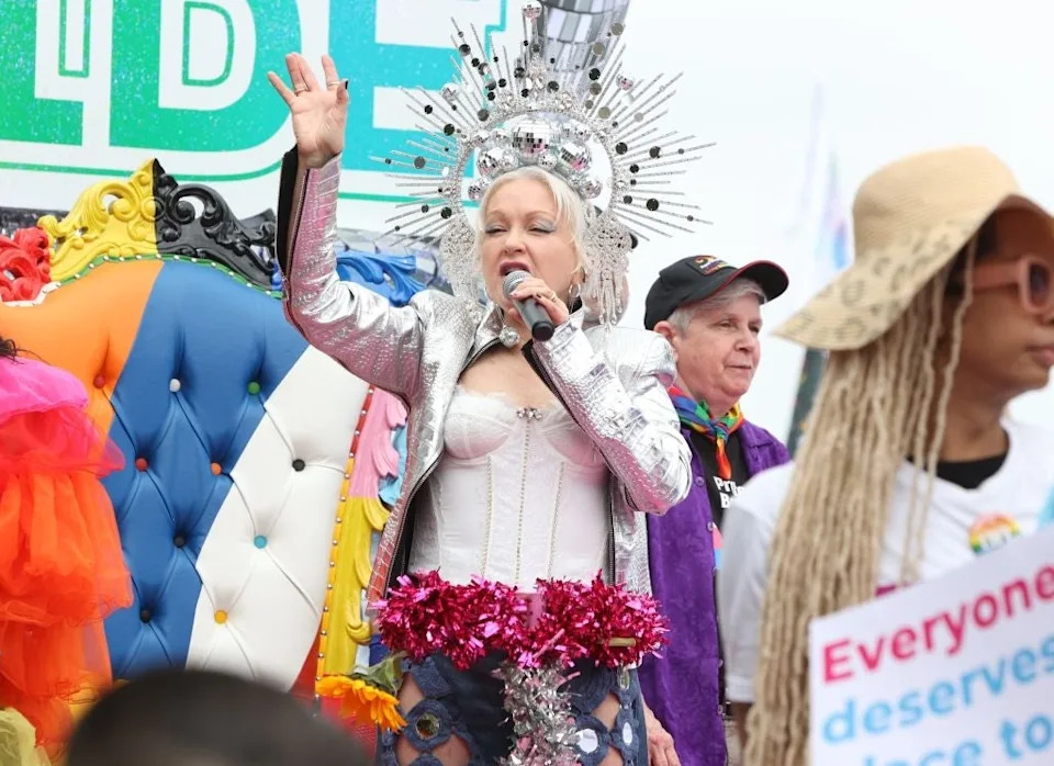 Person performing on stage wearing a metallic headdress and outfit with vibrant accents, holding a microphone. Several others are nearby