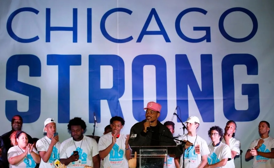 A person speaks at a podium in front of a large "Chicago Strong" sign, surrounded by people wearing matching shirts
