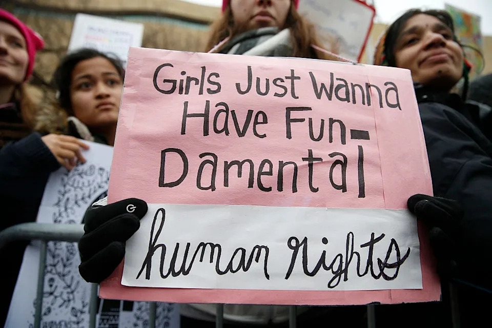 Protesters holding signs, including one reading "Girls Just Wanna Have Fundamental Human Rights" at a rally