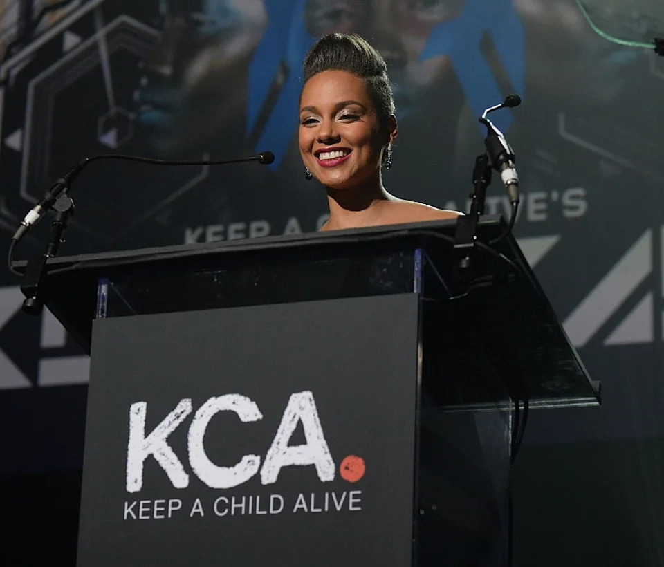 Person smiling at a podium with "Keep a Child Alive" text, likely at a fundraising or charity event