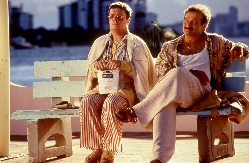 Two people sit on a bench; one wears a casual patterned shirt and the other wears a striped outfit. They're in a waterfront setting